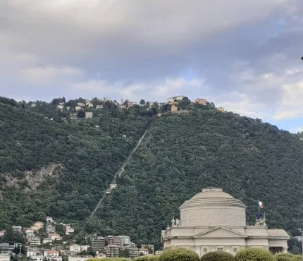 Inquadratura dal basso del Tempio Voltiano di Como con la sua cupola classica, sovrastato dalla verde collina che sale verso Brunate, lungo la quale si snoda la linea della funicolare.
